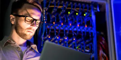 Ernster, beschäftigter junger IT-Ingenieur mit Brille steht vor Netzwerk-Server-Equipment mit leuchtenden Sensoren in einem dunklen Raum und nutzt einen Laptop, während er Netzwerksysteme überwacht.